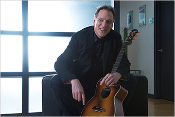 Image of FabCom's web developer Alfred smiling while wearing a black suit and holding an acoustic guitar.